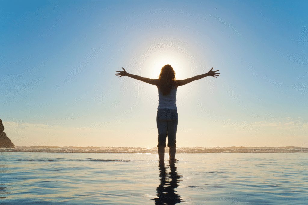 海辺の浅瀬で太陽に向かって両手を広げる女性の後ろ姿。自由と希望を感じさせる明るい風景