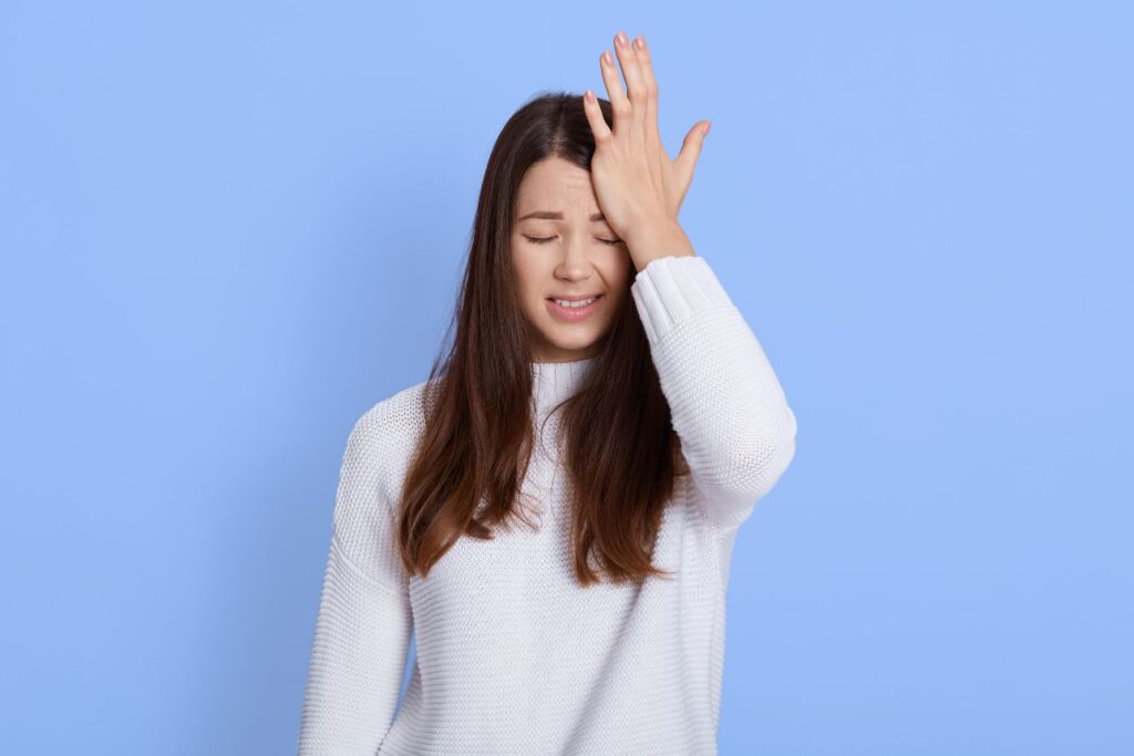 額に手を当てて目を閉じ、困った表情を浮かべる長い髪の若い女性。白いニットを着て、水色の背景の前に立っている。