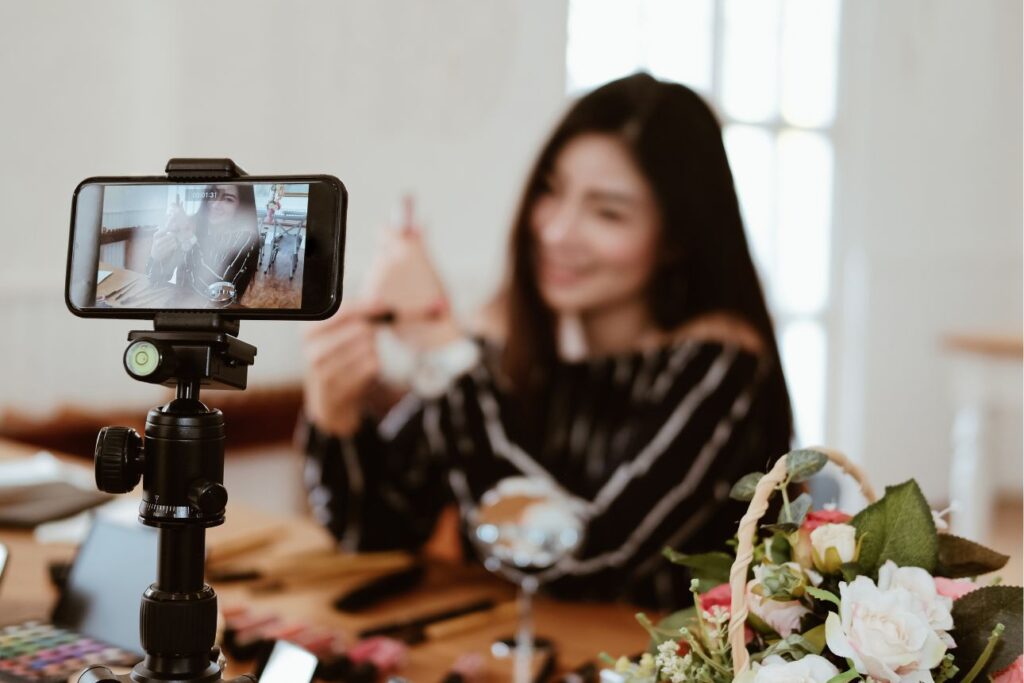 スマートフォンで自撮り動画を撮影する女性。化粧品を手に持ち、笑顔で話す様子がスマホ画面に映っている。