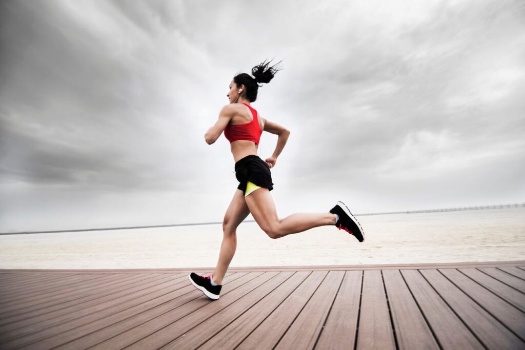 スポーツウェア姿の女性が海辺のボードウォークを全力でランニングしている様子。曇り空と開けた景色が背景。
