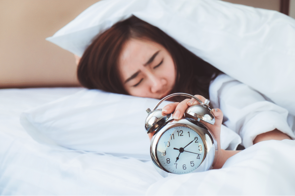 布団の中で目覚まし時計を手に持ち、眠そうな表情を浮かべる女性。睡眠不足や朝の倦怠感を表現