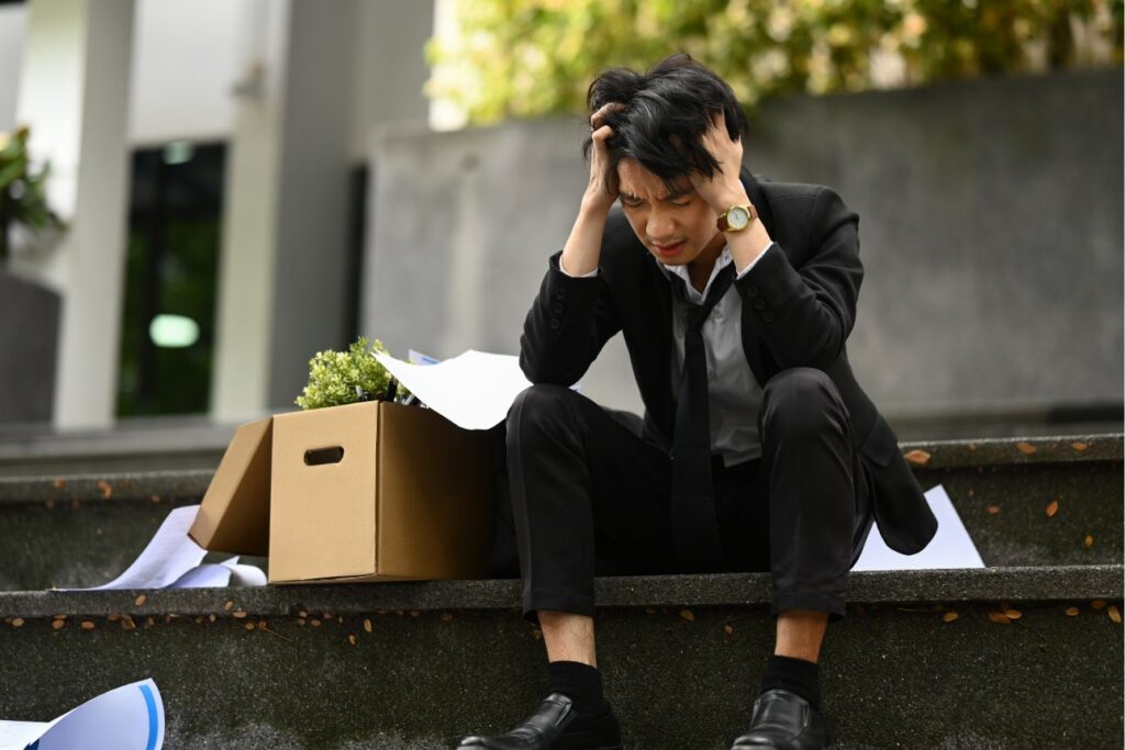 階段に座り頭を抱えて落ち込むスーツ姿の男性と荷物の入った段ボール箱