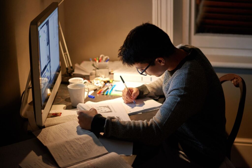 夜の部屋で机に向かい勉強する眼鏡の若い男性。