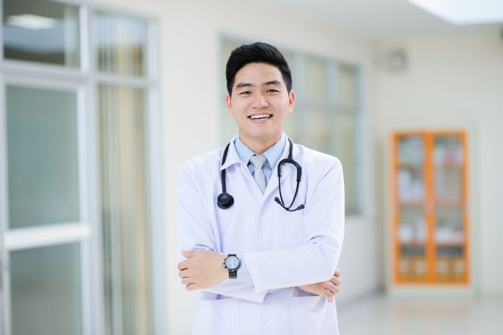 笑顔で腕を組む若い男性医師。白衣を着て聴診器を首にかけ、明るい医療施設内に立っている。