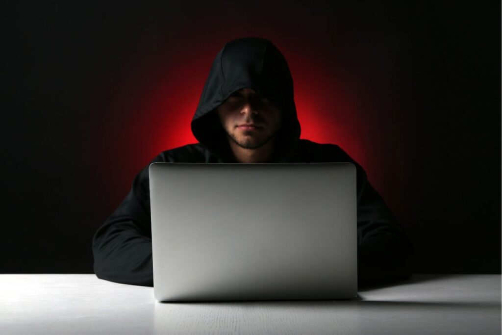 フードをかぶった人物がノートパソコンを操作する暗い室内の様子