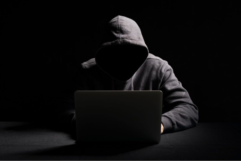 黒いフードをかぶった人物が暗闇の中でノートパソコンを操作している匿名性の高い姿