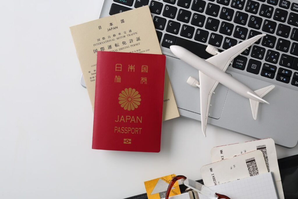 日本の赤いパスポートと国際運転免許証、ノートパソコンのキーボード、ミニチュア飛行機と搭乗券が揃った海外旅行の準備風景