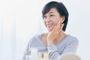 微笑みながら頬に手を添えるショートヘアの中年女性、明るい室内でスキンケアをする様子。