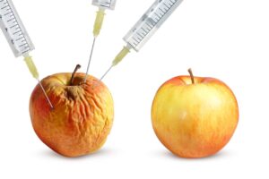 3本の注射器がしわしわに老化したリンゴに刺さっている様子と、その隣に置かれた若々しく新鮮なリンゴの対比。
