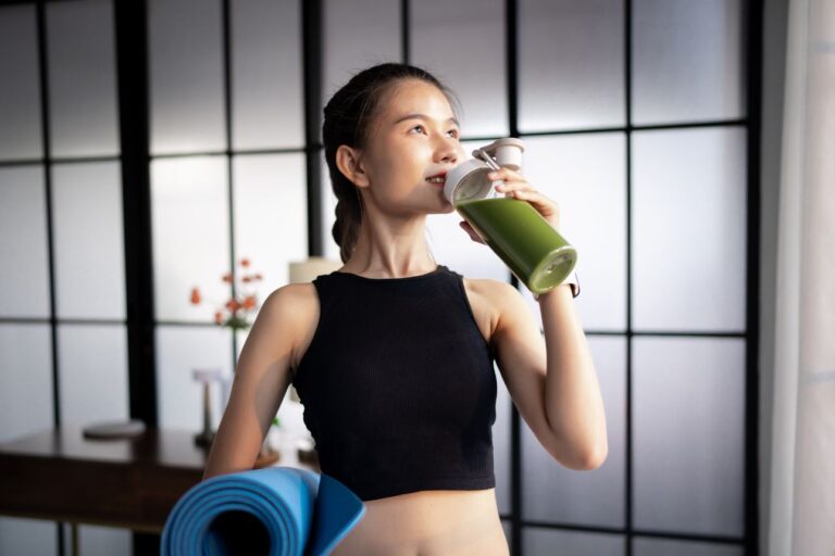 ヨガマットを持ち、グリーンスムージーを飲むフィットネスウェアの女性。ヘルシーなライフスタイルを象徴するシーン