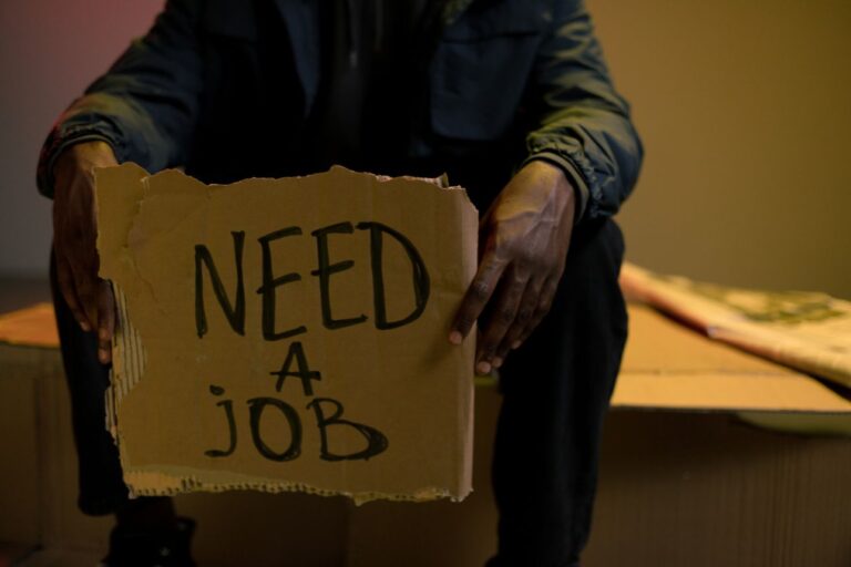 段ボールに「NEED A JOB」と書かれたサインを持つ人物の手元