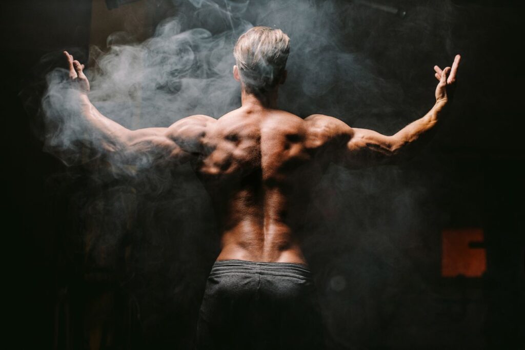 筋肉質な男性が背中を見せてポージングしている様子。煙が立ち込める中、力強い体が強調されている