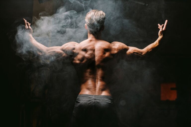 筋肉質な男性が背中を見せてポージングしている様子。煙が立ち込める中、力強い体が強調されている