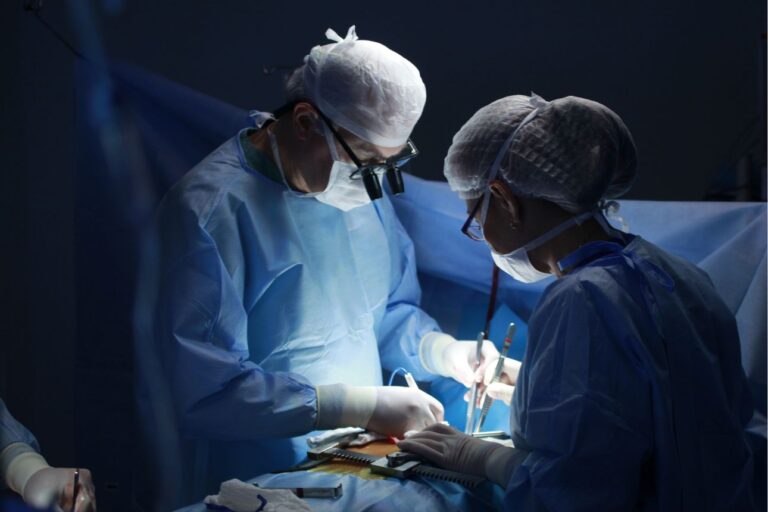 手術室で手術を行う2人の医師。青い手術着とマスク、拡大鏡を装着し、患者の患部を慎重に操作している様子。