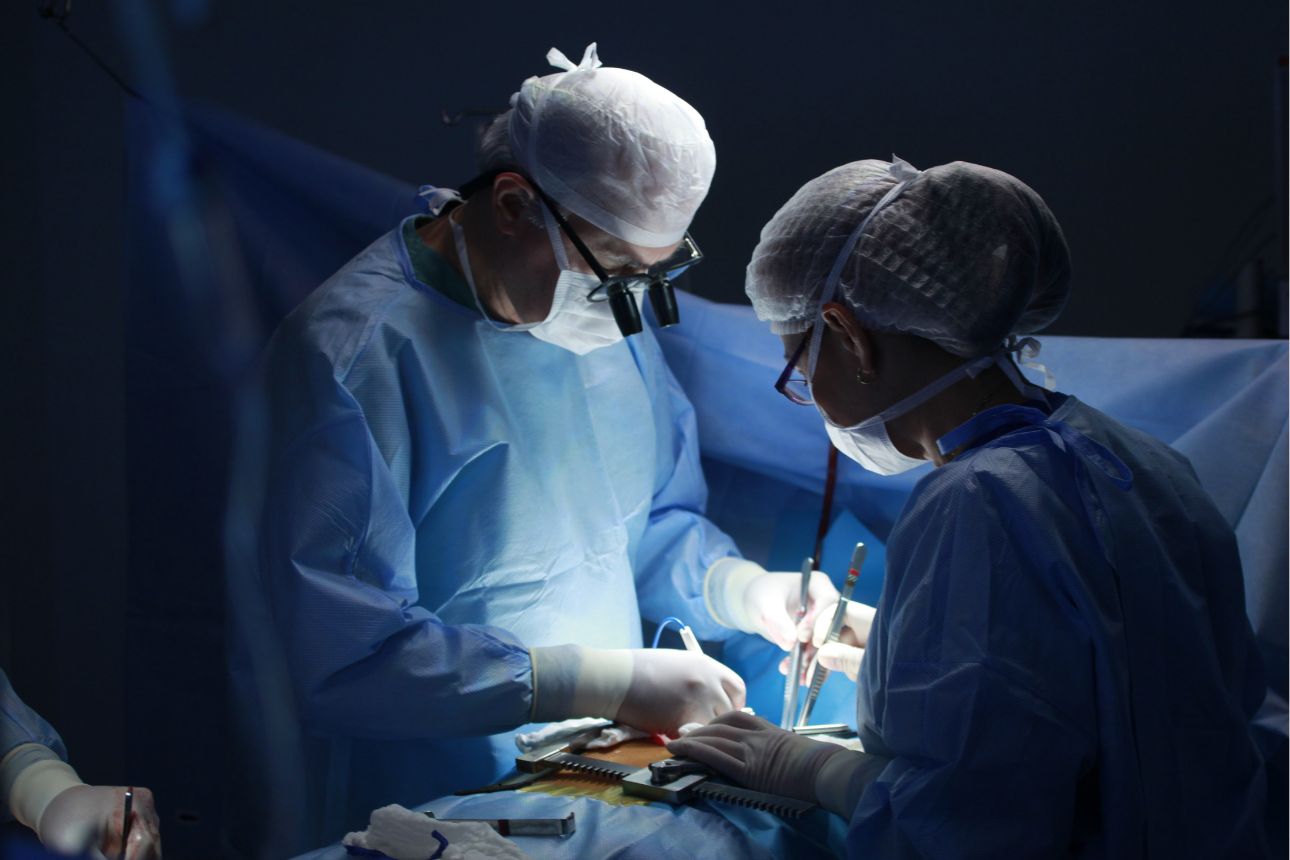 手術室で手術を行う2人の医師。青い手術着とマスク、拡大鏡を装着し、患者の患部を慎重に操作している様子。