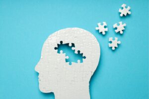 青い背景の上に置かれた人の横顔の形をした白いパズル。頭部の一部が欠けており、外に飛び出した数ピースのパズルが記憶の喪失や認知症を象徴している。