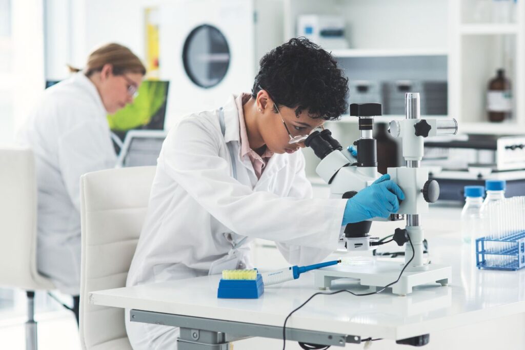顕微鏡でサンプルを観察する研究者と、背後でデータ解析を行う同僚研究員がいるバイオラボの様子
