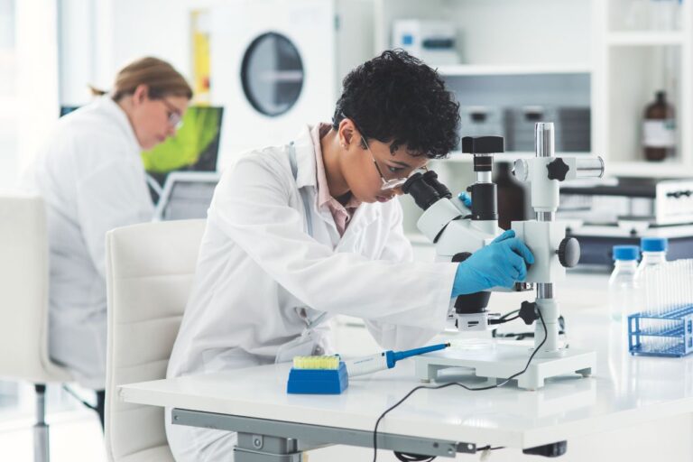 顕微鏡でサンプルを観察する研究者と、背後でデータ解析を行う同僚研究員がいるバイオラボの様子