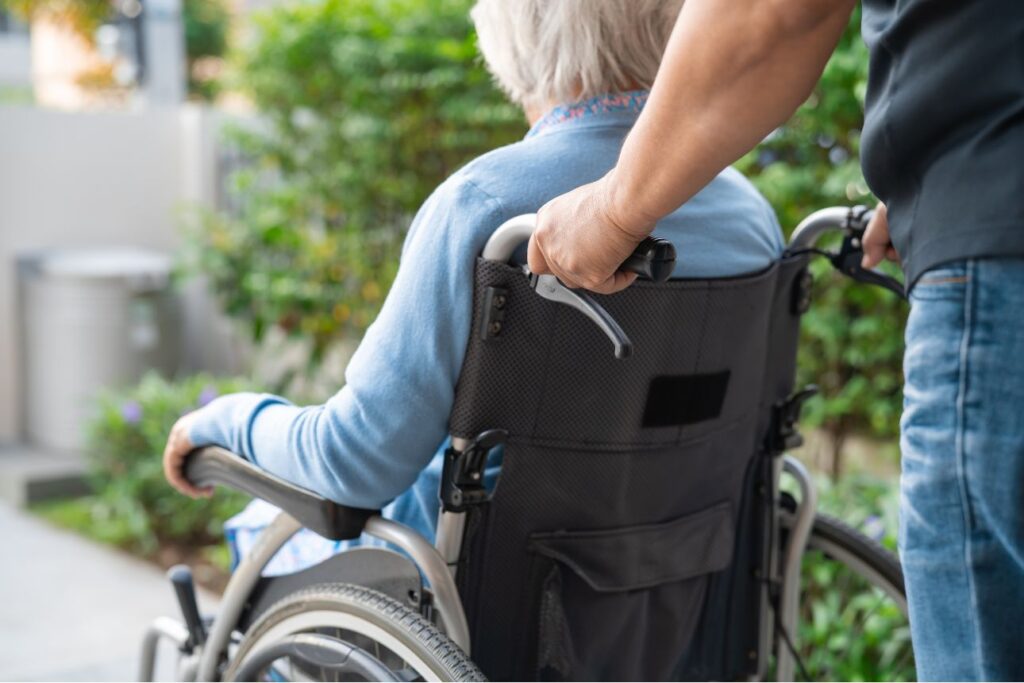 車椅子に座る高齢女性と、それを押す介助者の手元の様子。屋外の緑に囲まれた穏やかな風景