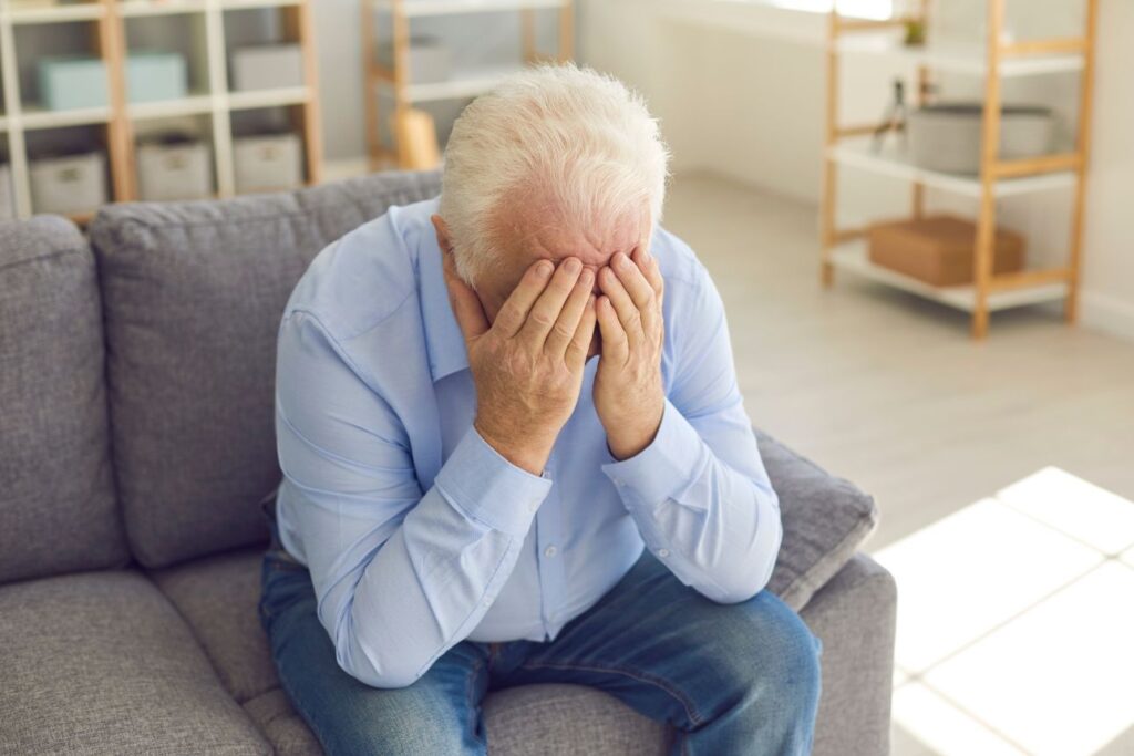 ソファに座り、両手で顔を覆って落ち込んでいる高齢の男性