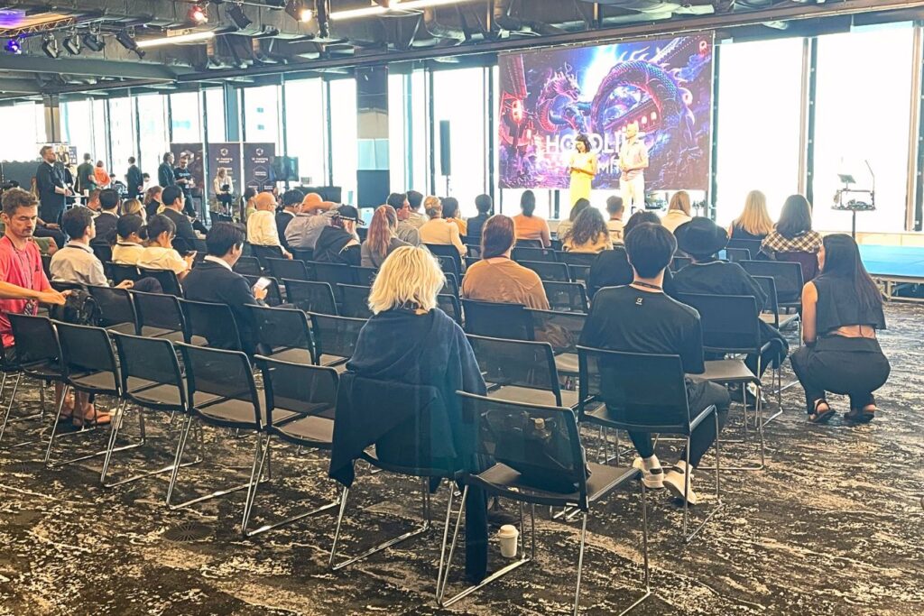 HOLOLIFE SUMMITの講演会場の様子。窓から自然光が差し込む渋谷の高層ビル内で、スクリーンには龍のビジュアルとイベントロゴが映し出され、登壇者2名がステージで話している。客席には国内外の参加者が座り、真剣に耳を傾けている。