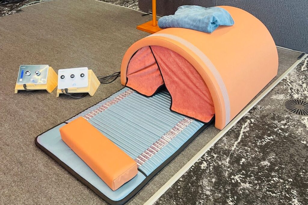 会場に設置された温熱ドーム型のウェルネス機器。オレンジ色の半円形ドームとマット、枕がセットになっており、横に温度や出力を調整するコントロールパネルが2台配置されている。リラクゼーションやバイオハッキング向けの体験設備として展示されている。